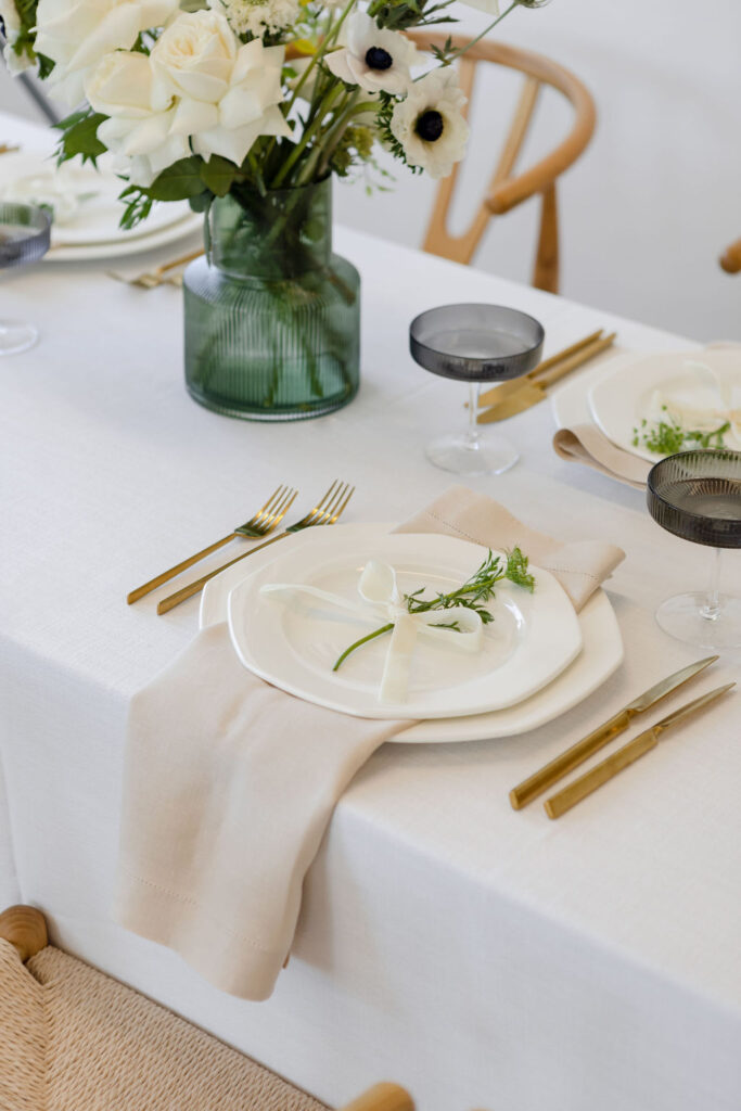 A Christmas table setting with white tablecloth & green glasses & white napkin on white plates with gold forks & knifes from hire society