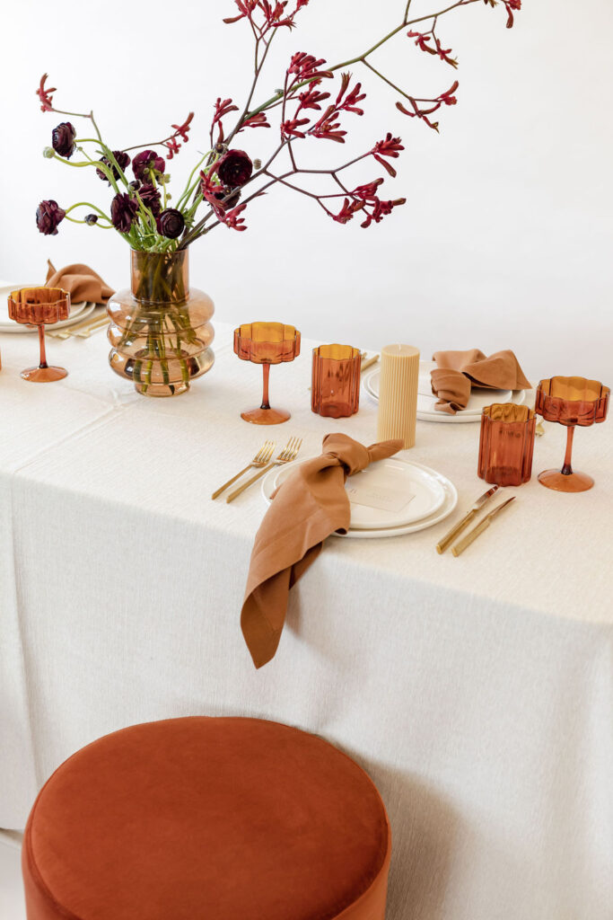 A Christmas table setting with white tablecloth & brown glasses & napkin on white plates with one brown chairs from hire society