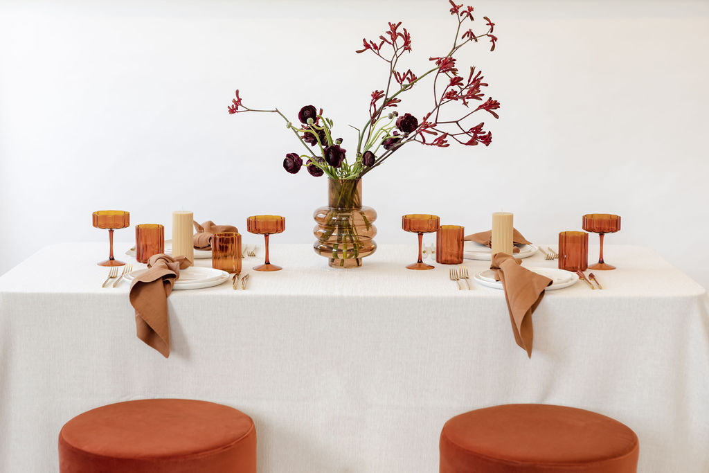 A Christmas table setting with white tablecloth and dark red flowers & orange glasses from hire society