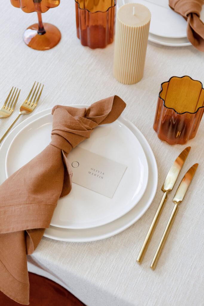 A Christmas table setting with white tablecloth & brown glasses & napkin on white plates from hire society